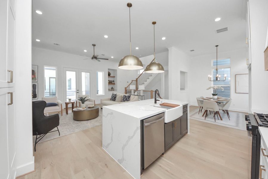 This bright, modern living room radiates warmth and style. Light wood floors and a ceiling fan complement the abundance of natural light streaming through large windows. A comfortable seating area with neutral tones, decorative pillows, and stylish accents creates an inviting atmosphere, while contemporary art, a sleek staircase railing, and indoor plants add touches of sophistication and greenery.