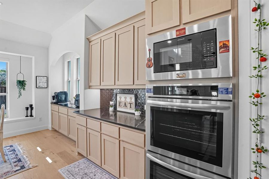 Kitchen featuring appliances with stainless steel finishes, light wood-type flooring, light brown cabinetry, dark stone counters, and backsplash