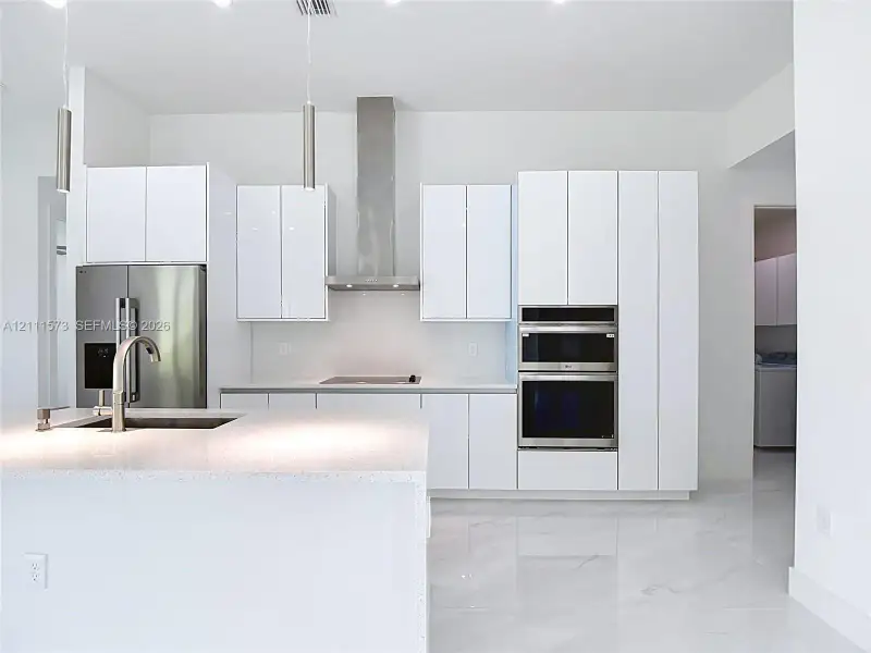 Kitchen featuring white high-gloss cabinetry, stainless steel built-in double ovens and range hood, center island with stone-surface countertop, and large-format porcelain floor tile