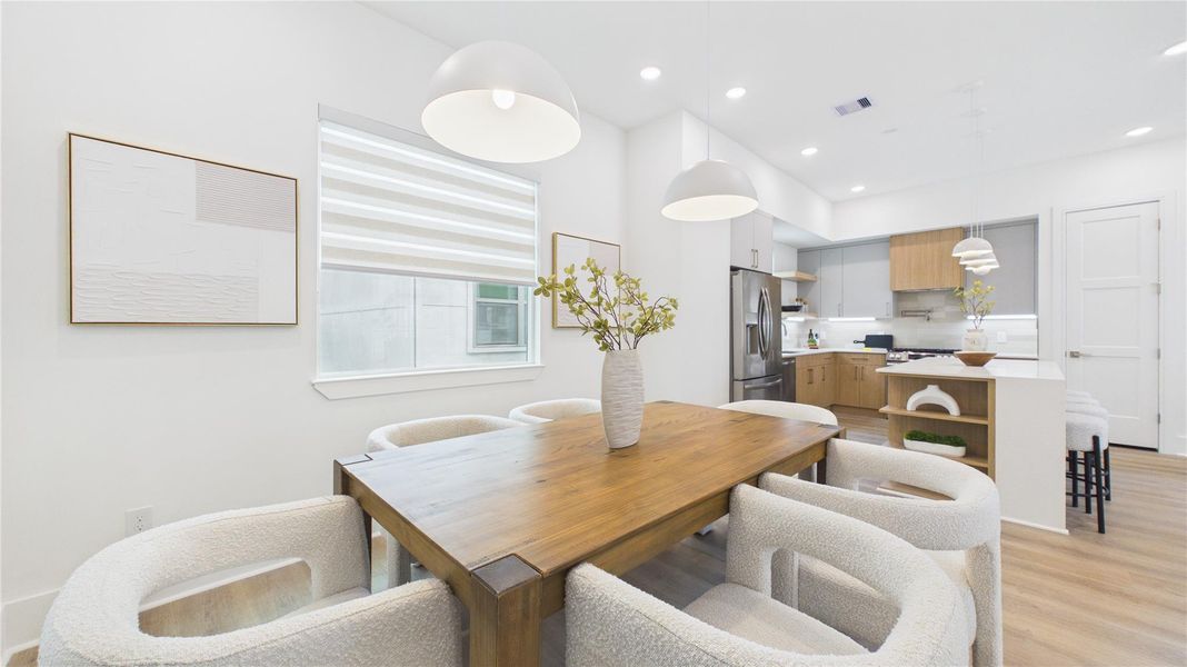 This modern, open-concept kitchen and dining area features a sleek design with white walls, wooden floors, and contemporary pendant lighting. The kitchen includes stainless steel appliances and a cozy island with seating. The dining area boasts a stylish wooden table and comfortable chairs, creating a welcoming space for meals and gatherings.