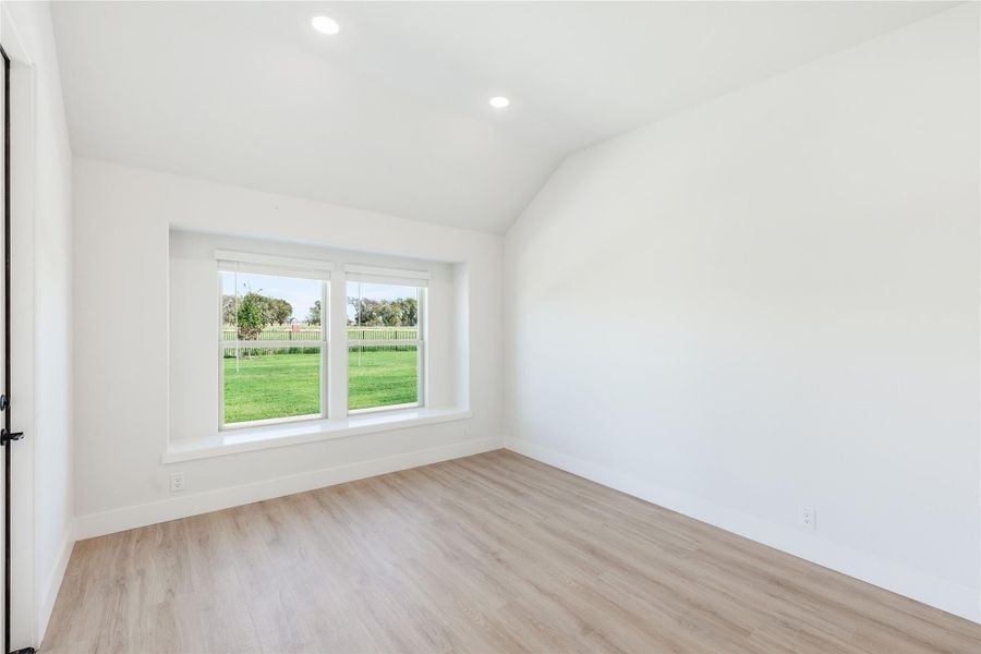Empty room with vaulted ceiling, light wood floors, and double window overlooking green yard