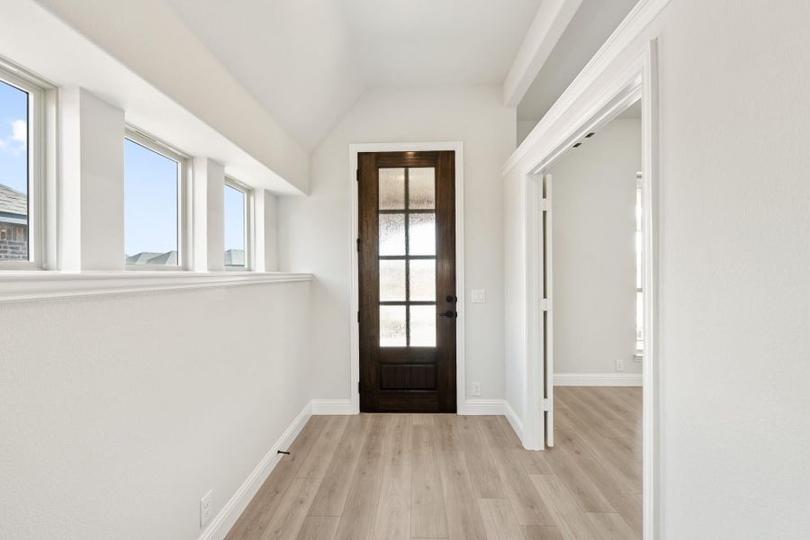 Entryway hallway with dark wood glass-panel door, light hardwood floors, and row of windows along white walls Entryway hallway with dark wood glass-panel door, light hardwood floors, and row of windows along white walls