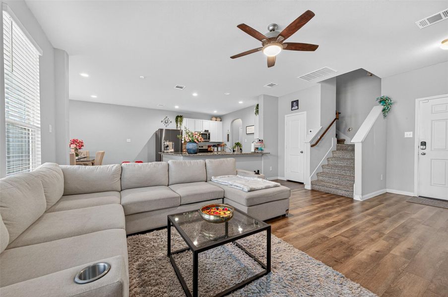 This spacious living area features an open floor plan with a large sectional sofa, modern ceiling fan, and hardwood floors. The kitchen and dining area are visible in the background, providing a seamless flow. There's also a staircase leading to the upper level, adding to the home's inviting and functional layout.