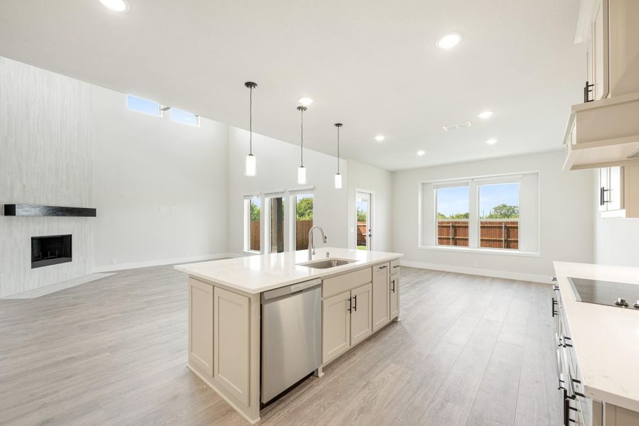 Open kitchen with center island, pendant lights, and stainless dishwasher, open to living room with fireplace and hardwood floors