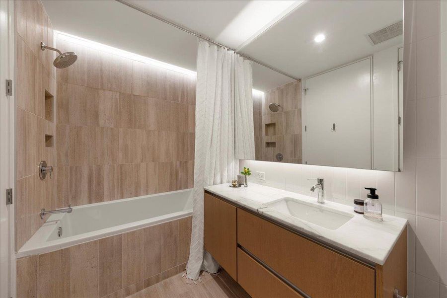 Contemporary second bath featuring a sleek double vanity with custom marble counters, modern fixtures and a soaking tub/shower combination wrapped in warm, textured tile. A refined and functional space designed for guests or everyday living.
