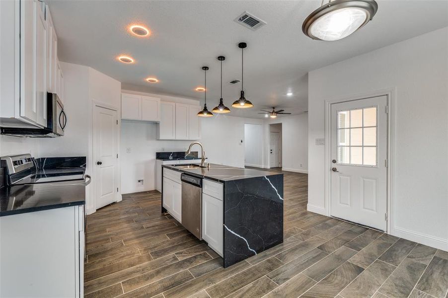 Kitchen featuring wood tiled floors, a kitchen island with sink, decorative light fixtures, white cabinetry, and appliances with stainless steel finishes Kitchen featuring wood tiled floors, a kitchen island with sink, decorative light fixtures, white cabinetry, and appliances with stainless steel finishes