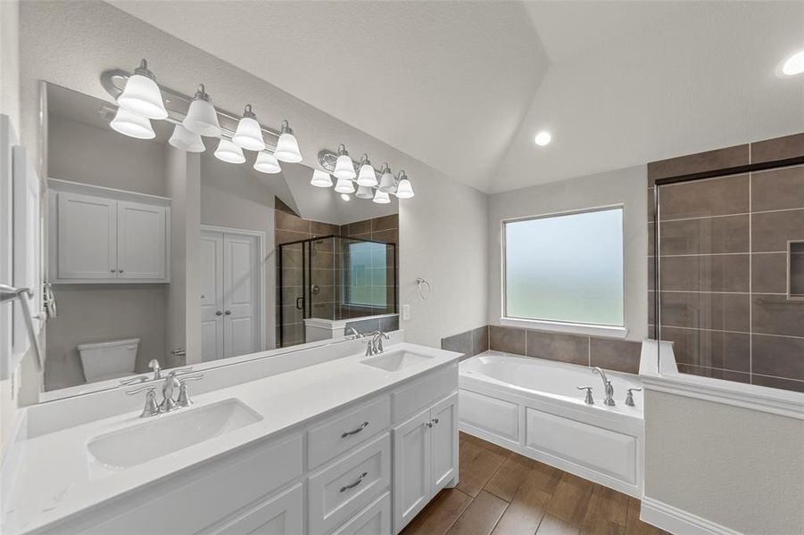 Spacious bathroom featuring a dual vanity with white cabinetry and chrome fixtures, a large soaking tub, a tiled walk-in shower with a glass enclosure, and wood-finish flooring
