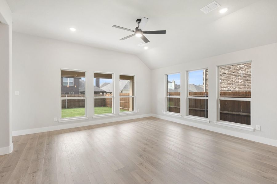 Empty living room with vaulted ceiling, ceiling fan, light wood floors, and large windows overlooking backyard