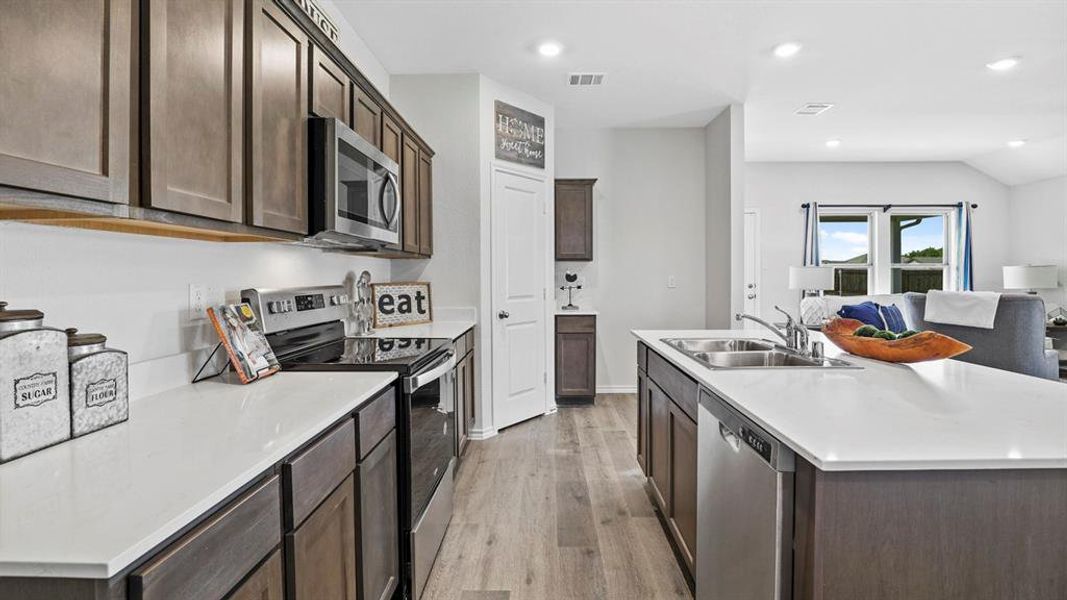 Contemporary kitchen featuring wood-finish flooring, stainless steel appliances, white countertops, dark wood cabinetry, and an island with an undermount double-basin sink
