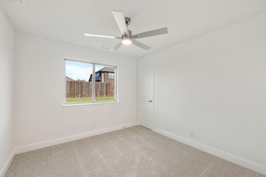 Empty bedroom with white walls, carpet flooring, ceiling fan, and a window overlooking a backyard fence