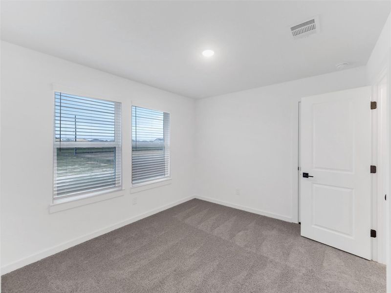 Secondary bedroom with neutral color carpeting and two windows allowing natural light. The walls are painted white, and there's a closed door, making it a versatile space for a bedroom or office.