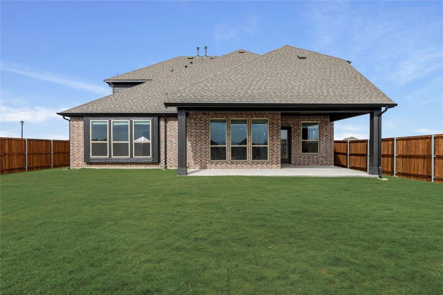 Rear view of brick home with covered patio, large windows, and fenced backyard with green lawn Rear view of brick home with covered patio, large windows, and fenced backyard with green lawn
