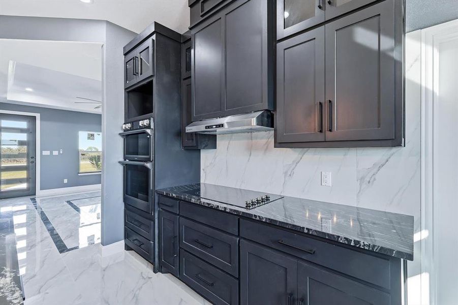 Kitchen with light marble finish floors, dark stone countertops, stainless steel double oven, under cabinet range hood, and decorative backsplash
