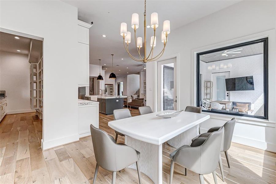 This bright breakfast area features a modern table, stylish chairs, and a chic chandelier. It opens to a spacious kitchen with pendant lighting and offers views of a cozy outdoor patio through large windows.