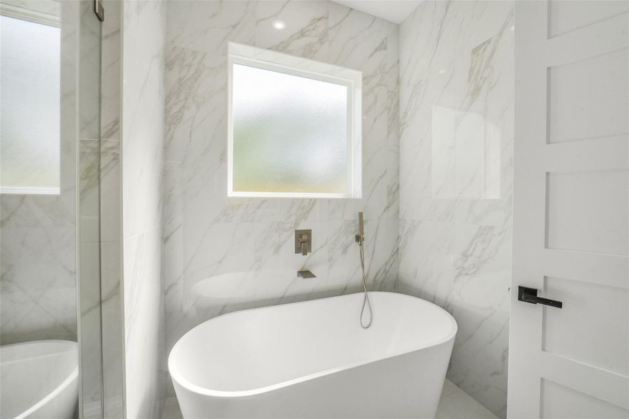 This bathroom features a modern design with a freestanding tub and elegant marble-patterned walls. It includes a frosted window for natural light and privacy, creating a bright and inviting space.