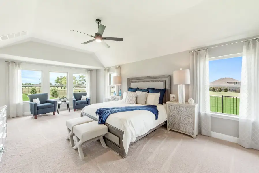 Spacious master bedroom with vaulted ceiling, ceiling fan, large windows, and a seating area with two blue chairs.