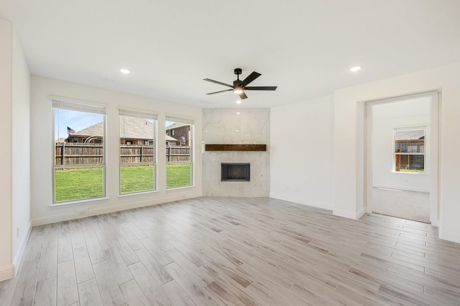 Open living room with marble fireplace, ceiling fan, light wood floors, and large windows overlooking backyard Open living room with marble fireplace, ceiling fan, light wood floors, and large windows overlooking backyard