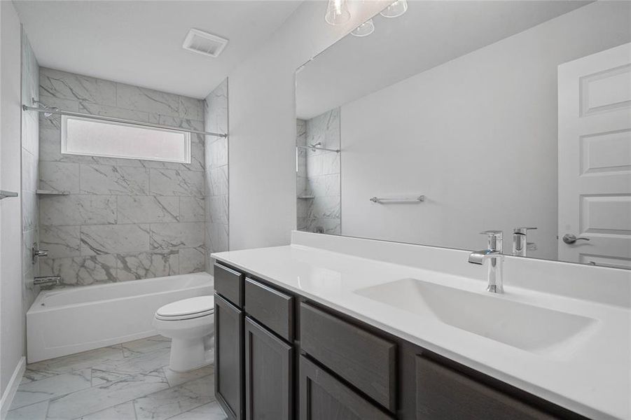 Secondary bath features tile flooring, bath/shower combo with tile surround, stained wood cabinets, beautiful light countertops, mirror, dark, sleek fixtures and modern finishes.