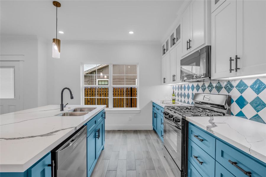 This kitchen boasts a modern flair with its striking blue cabinetry, sleek stainless steel appliances, and elegant marble countertops, all illuminated by stylish pendant lighting for a bright, inviting atmosphere.