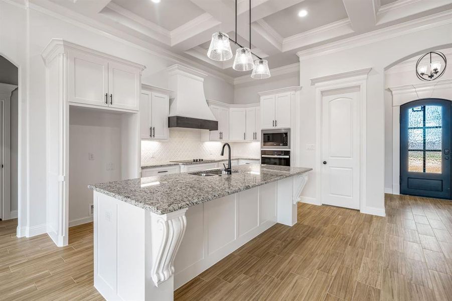 Kitchen with custom exhaust hood, light stone countertops, built in microwave, an island with sink, and white cabinets