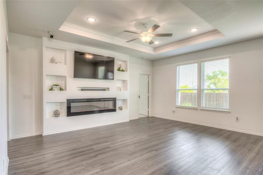 Unfurnished living room with ornamental molding, a glass covered fireplace, wood finished floors, a tray ceiling, and ceiling fan Unfurnished living room with ornamental molding, a glass covered fireplace, wood finished floors, a tray ceiling, and ceiling fan