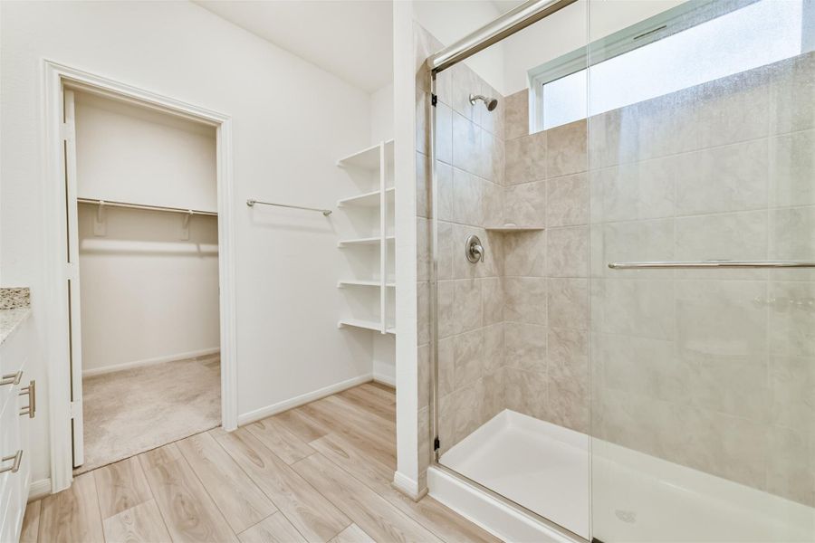 Another view of the spacious walk-in shower with a glass door and modern tile in the Primary bathroom. It includes built-in shelving, a granite countertop, and access to a large walk-in closet. The room is bright with neutral tones and wood flooring.