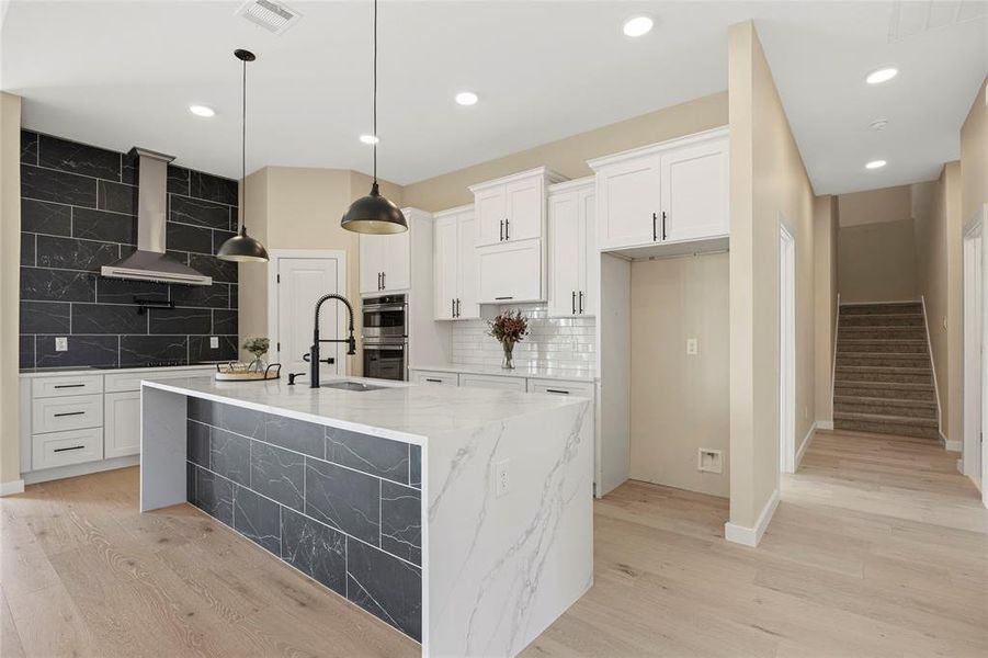 Kitchen featuring wall chimney range hood, stainless steel double oven, white cabinetry, light stone counters, and a kitchen island with sink