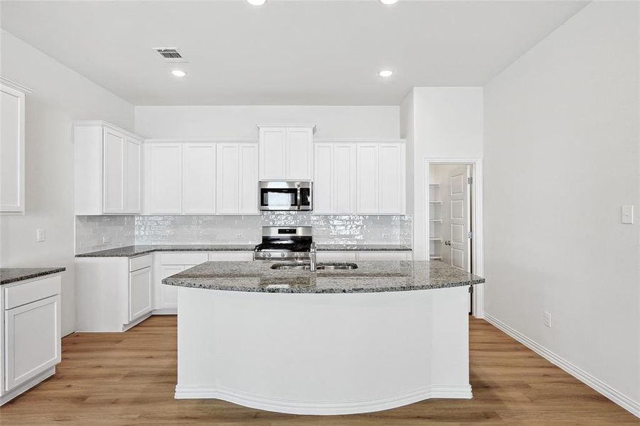 Kitchen featuring dark stone counters, light hardwood / wood-style flooring, stainless steel appliances, tasteful backsplash, and a kitchen island with sink