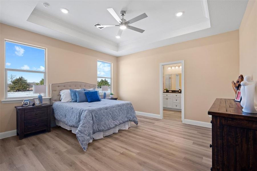 Spacious bedroom featuring light-toned walls, wood-finish flooring, and a tray ceiling with recessed lighting and a ceiling fan Spacious bedroom featuring light-toned walls, wood-finish flooring, and a tray ceiling with recessed lighting and a ceiling fan