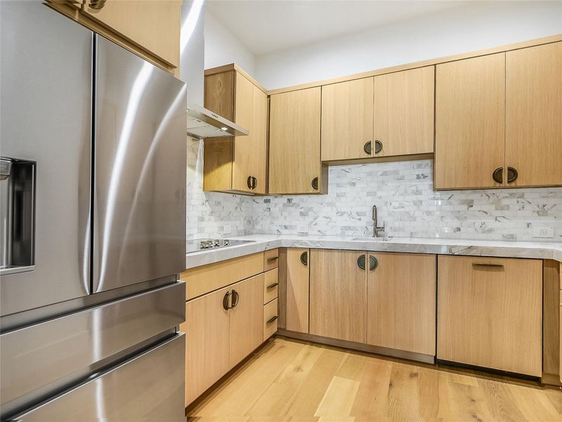 The kitchen features natural wood cabinetry, a stainless steel refrigerator, white marble countertops, and a white tile backsplash