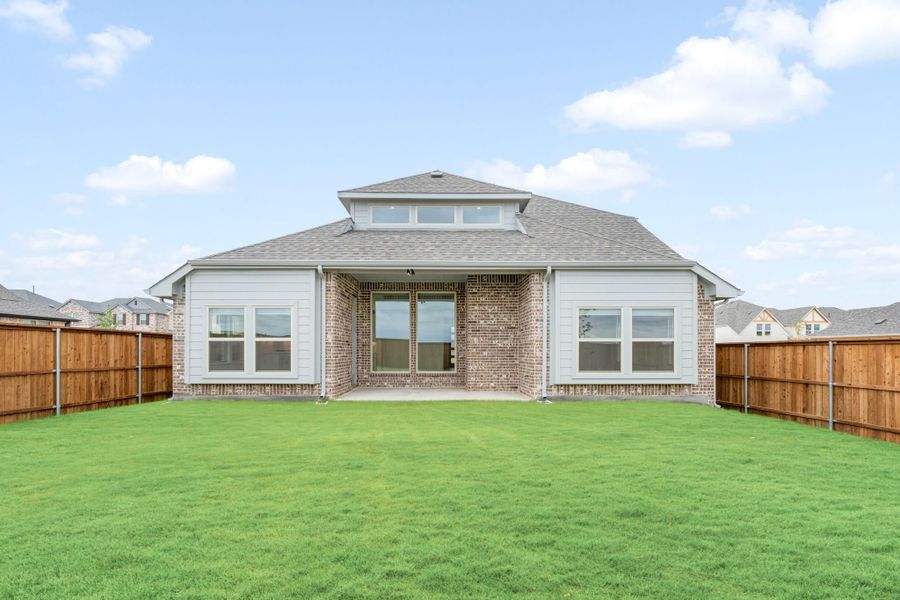 Rear view of single-story brick home with covered patio, large green backyard, and wood privacy fence