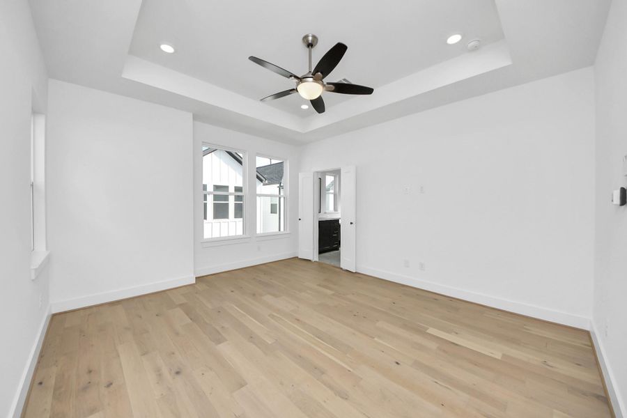 This bright, spacious room features light hardwood flooring, a tray ceiling with recessed lighting, and a ceiling fan. Large windows provide ample natural light, and the room offers access to an adjoining space or bathroom through a doorway.