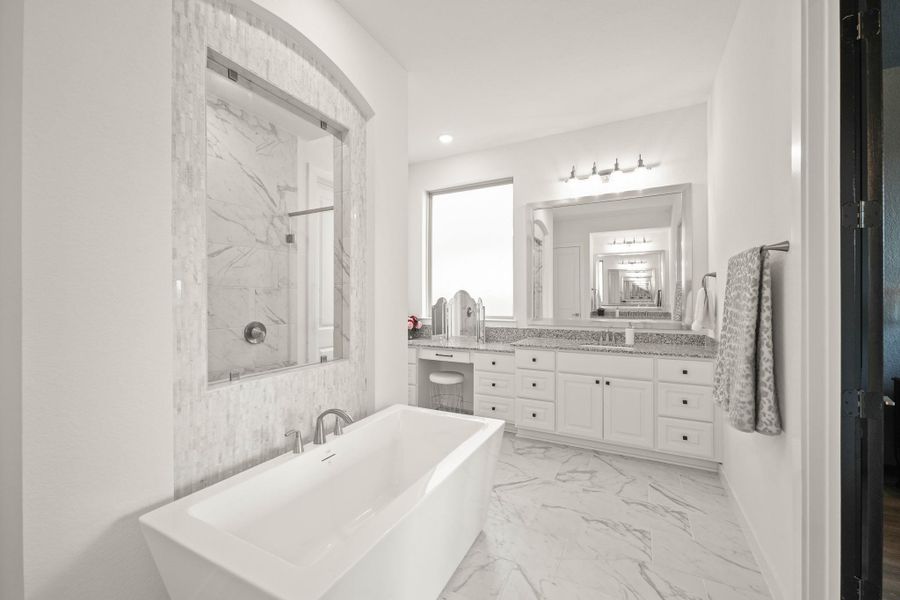 A freestanding soaking tub with a satin nickel faucet and a tiled accent wall adds a spa-like touch to the space, while the granite-surfaced counter with under mount sink and separate make-up vanity area includes a fixed window with opaque glass. The framed mirrors with lighting above illuminate the room.