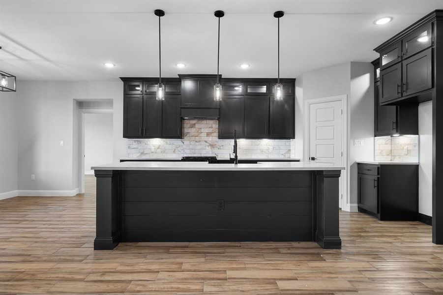 Kitchen featuring decorative backsplash, a center island with sink, hanging light fixtures, and range hood