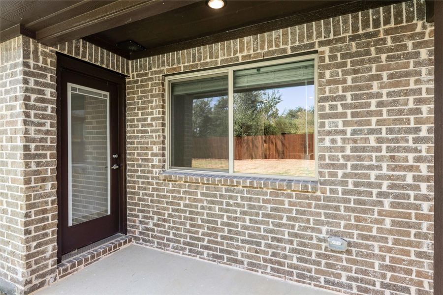 Covered back patio with brick exterior wall, dark wood door, and sliding window overlooking fenced backyard