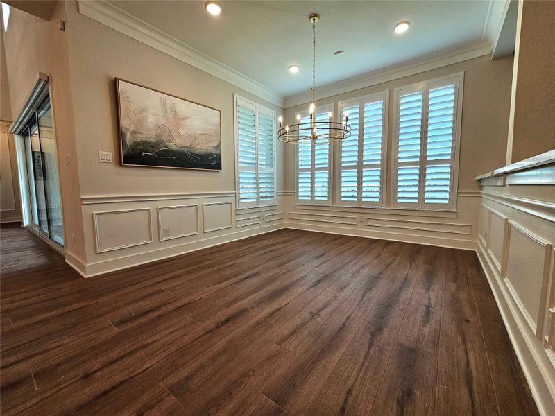 This room features elegant wood flooring, large windows with plantation shutters, and stylish wainscoting. A modern chandelier adds a touch of sophistication, making it perfect for a dining or living area.