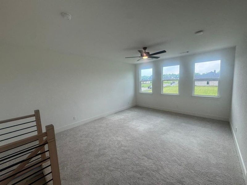 Spacious carpeted room featuring a ceiling fan, three large windows, and a contemporary wood and metal railing