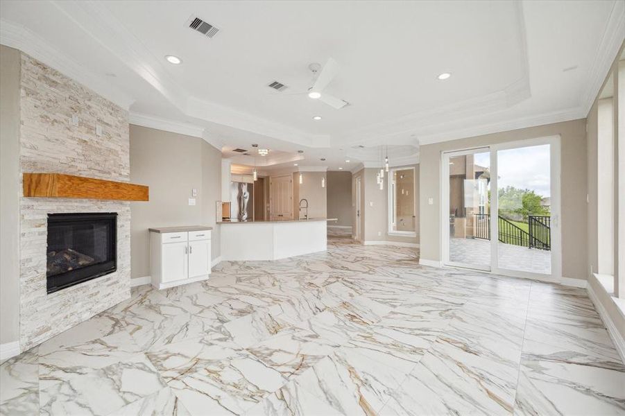 This image showcases a spacious, modern living area with high ceilings and marble flooring. The room features a stone-clad fireplace, a kitchen with a bar counter in the background, and large sliding doors leading to an outdoor balcony with a railing.