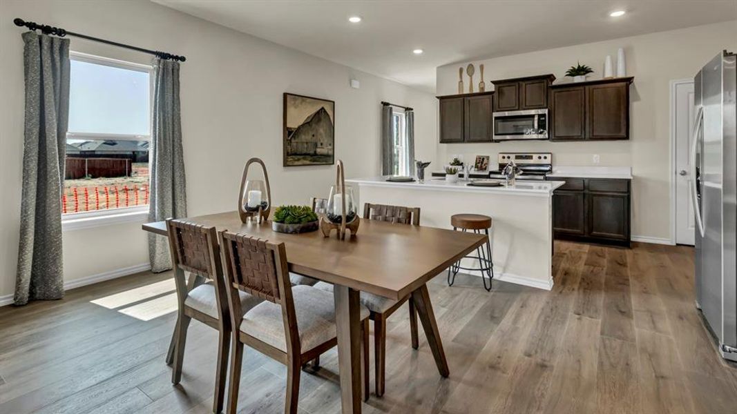 Open-concept dining area featuring wood-finish flooring, a contemporary window with drapes, a kitchen with dark cabinetry, stainless steel appliances, and a breakfast bar with a white countertop Open-concept dining area featuring wood-finish flooring, a contemporary window with drapes, a kitchen with dark cabinetry, stainless steel appliances, and a breakfast bar with a white countertop