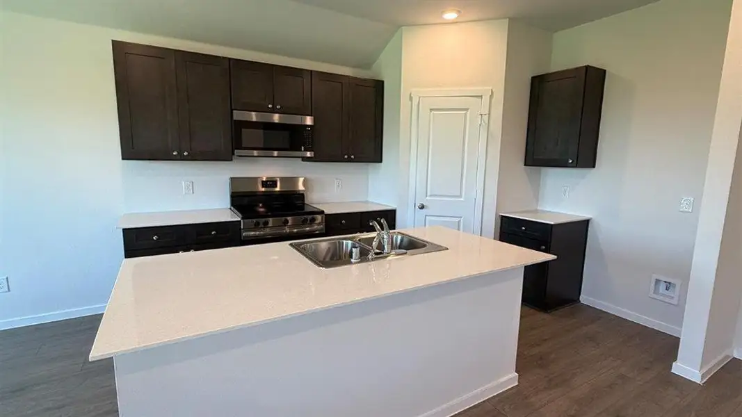 Kitchen featuring a center island with double basin sink, dark wood cabinetry, stainless steel range with overhead microwave, light-toned countertops, and wood-finish flooring Kitchen featuring a center island with double basin sink, dark wood cabinetry, stainless steel range with overhead microwave, light-toned countertops, and wood-finish flooring