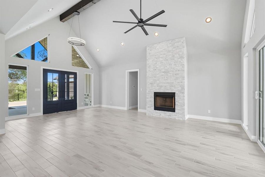 Unfurnished living room with high vaulted ceiling, french doors, beamed ceiling, light wood finished floors, and a stone fireplace Unfurnished living room with high vaulted ceiling, french doors, beamed ceiling, light wood finished floors, and a stone fireplace