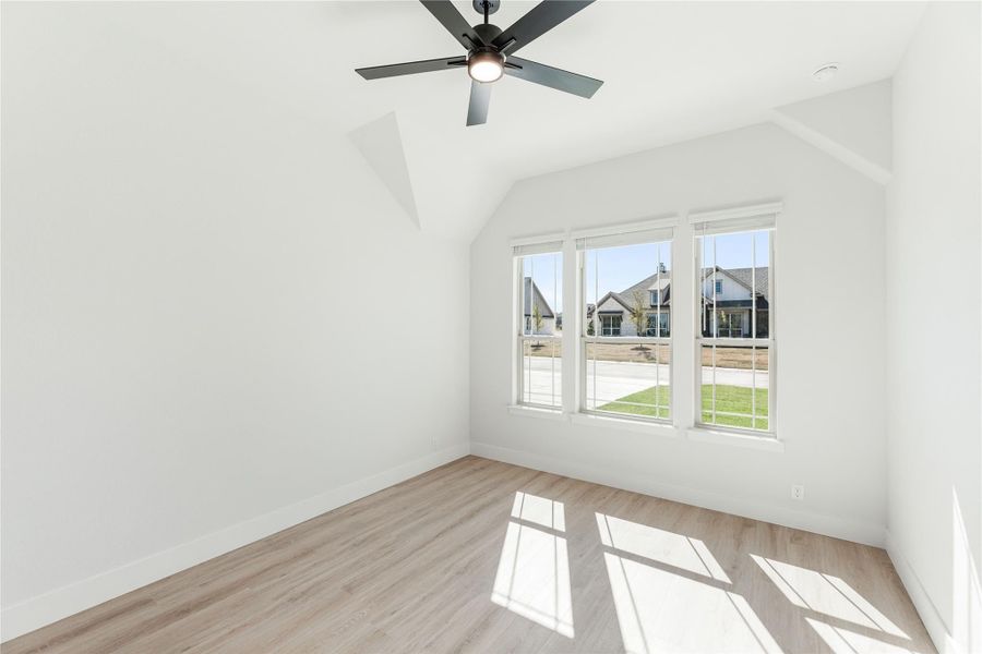 Empty bedroom with vaulted ceiling, ceiling fan, light wood floors, and triple windows overlooking neighborhood