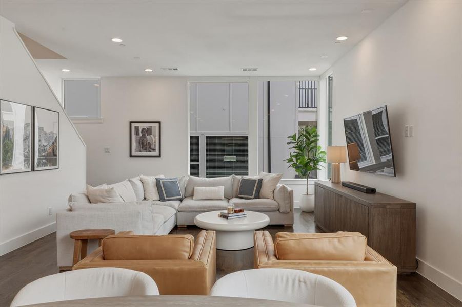 The living area features recessed lighting, large windows providing natural light, and dark hardwood flooring