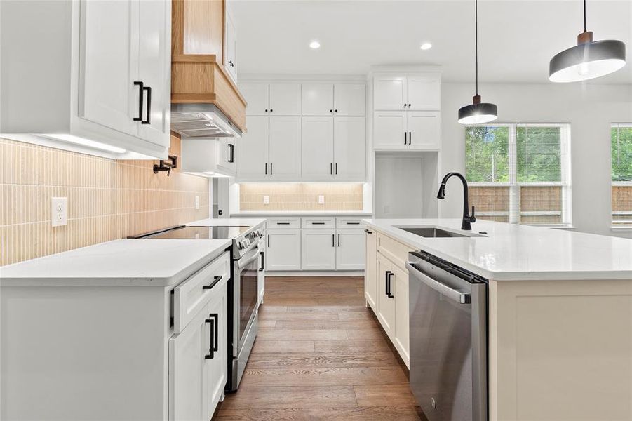 Open-concept kitchen featuring white shaker cabinetry, an expansive central island with an undermount sink, stainless steel appliances, wood-finish flooring, and a light-toned vertical tile backsplash