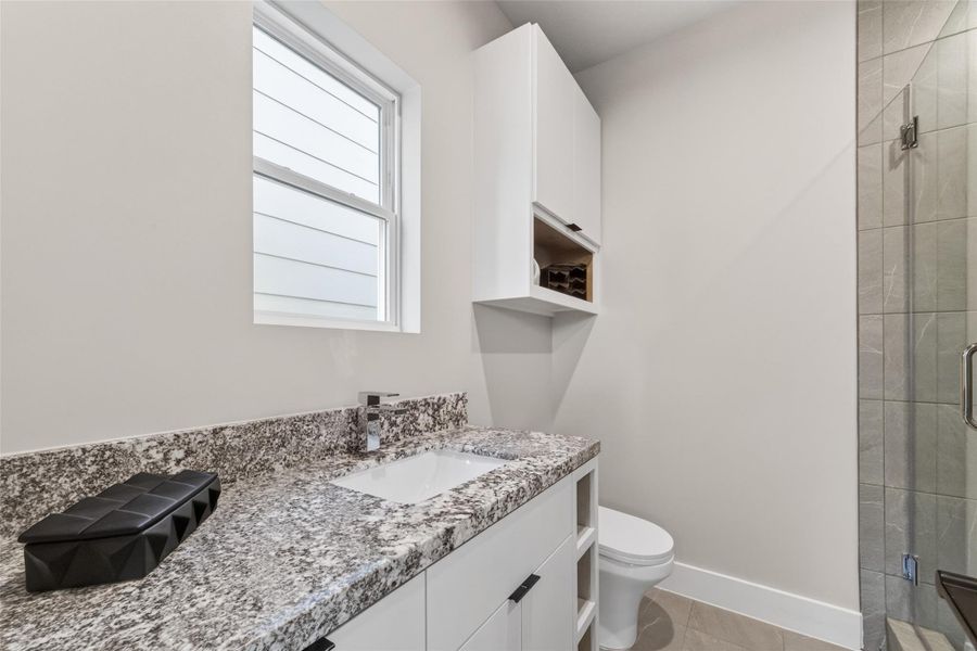 The third-floor secondary bath features a stone vanity countertop, tile walk-in shower with glass enclosure, and built-in overhead cabinetry. A bright window and clean white finishes give the room a fresh, modern feel.