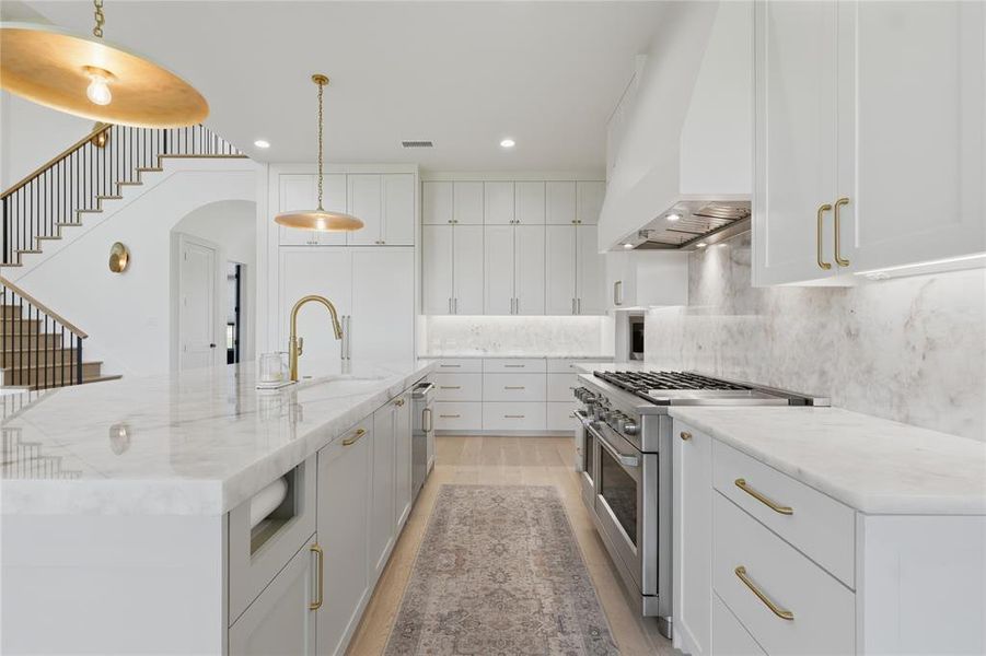 Gourmet kitchen featuring a substantial island with integrated sink, white cabinetry with gold-tone hardware, stainless steel range with double ovens, stone countertops and backsplash, and light wood-finish flooring