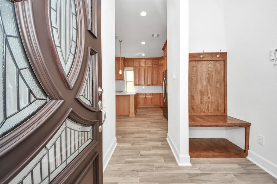 Welcome to your new home! This inviting entry features a stylish front door with decorative glass, leading to a bright interior with ceramic wood-look tile flooring. The entryway includes a convenient built-in bench with hooks for storage. The open view to the kitchen showcases beautiful wood cabinetry and modern finishes.