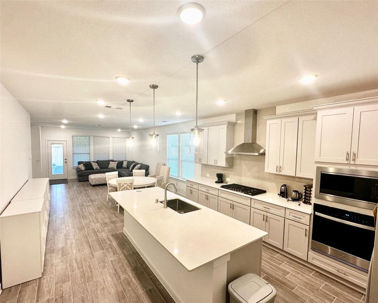 This open-concept space features a modern kitchen with an island, dining area, and living room. It has wood-look tile flooring, bright lighting, and a neutral color palette, creating a spacious and inviting atmosphere.