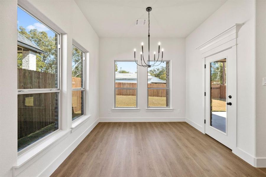 Bright interior space featuring wood-finish flooring, multiple windows, a ceiling-mounted chandelier, and a glass-paned door with decorative trim Bright interior space featuring wood-finish flooring, multiple windows, a ceiling-mounted chandelier, and a glass-paned door with decorative trim