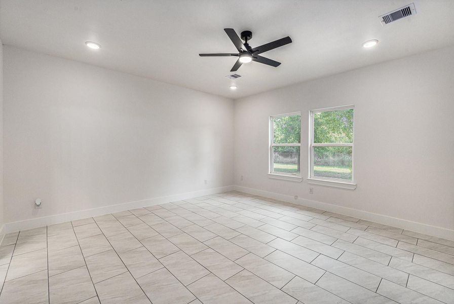 This room features a modern design with light-colored tile flooring, neutral walls, recessed lighting, and a ceiling fan. Two large windows offer plenty of natural light and a view of greenery outside.